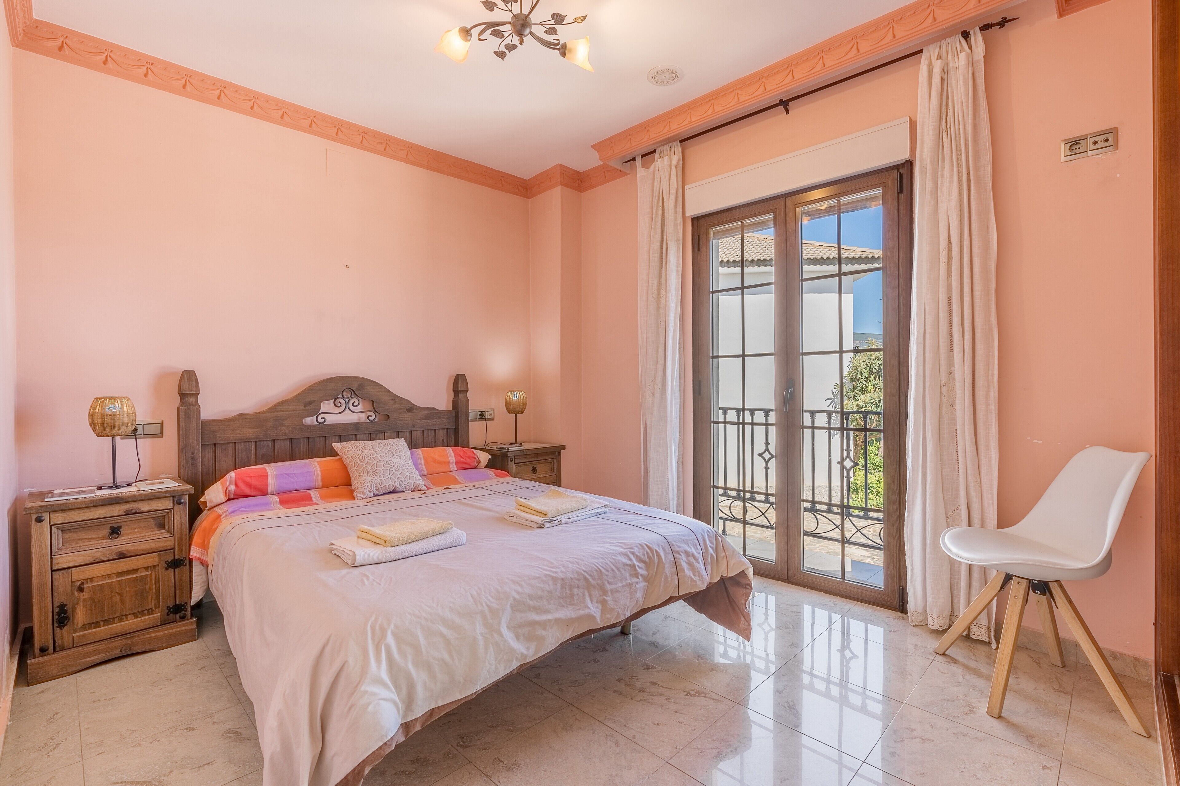 Une chambre à coucher comprenant un lit, des tables de chevet, une chaise, une fenêtre avec des rideaux et un balcon.