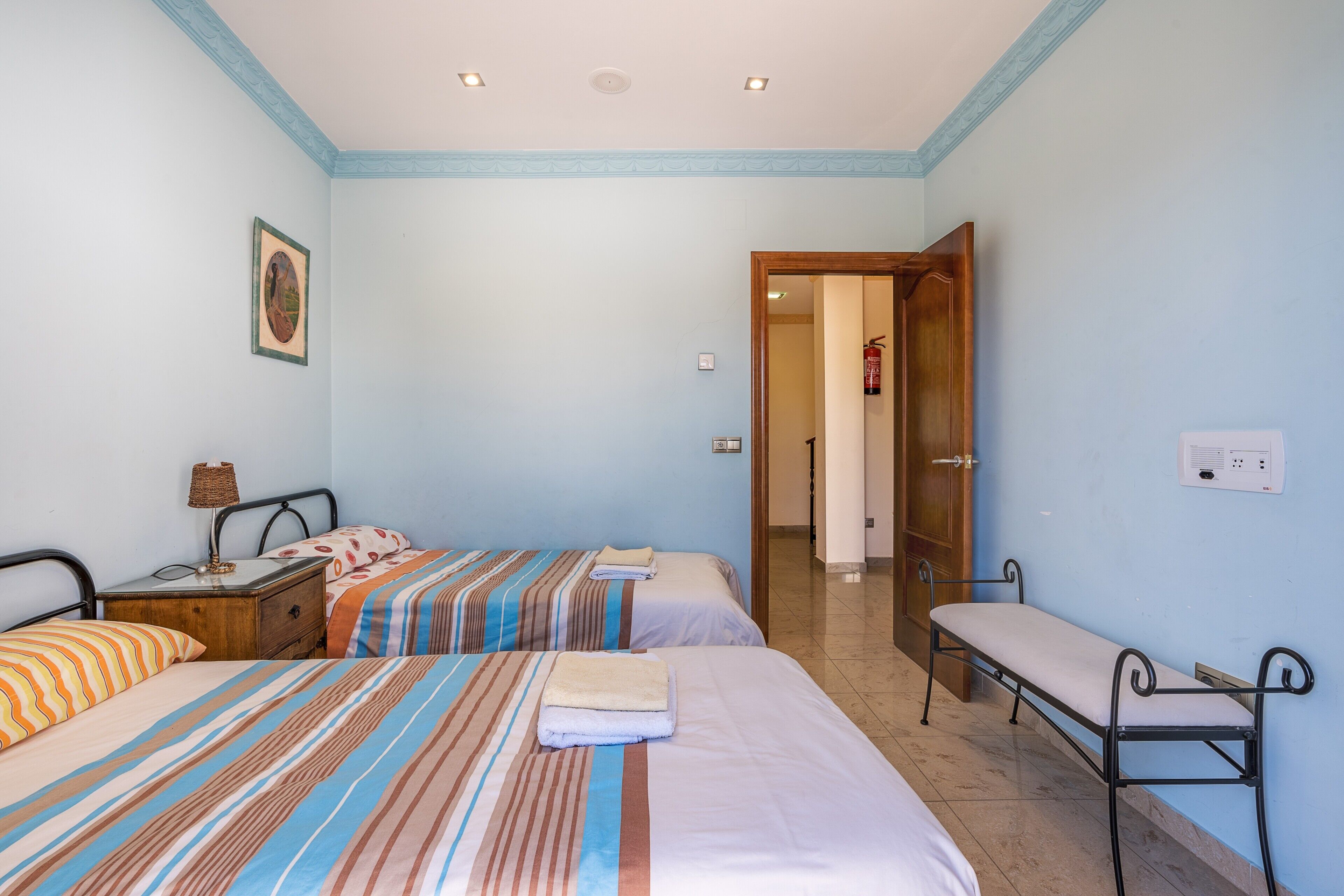 A room with two beds, a bench, a door, and a wall-mounted light fixture.