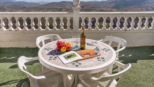 Una mesa redonda con un libro, una botella y fruta encima, rodeada de sillas blancas, con vistas a montañas al fondo.