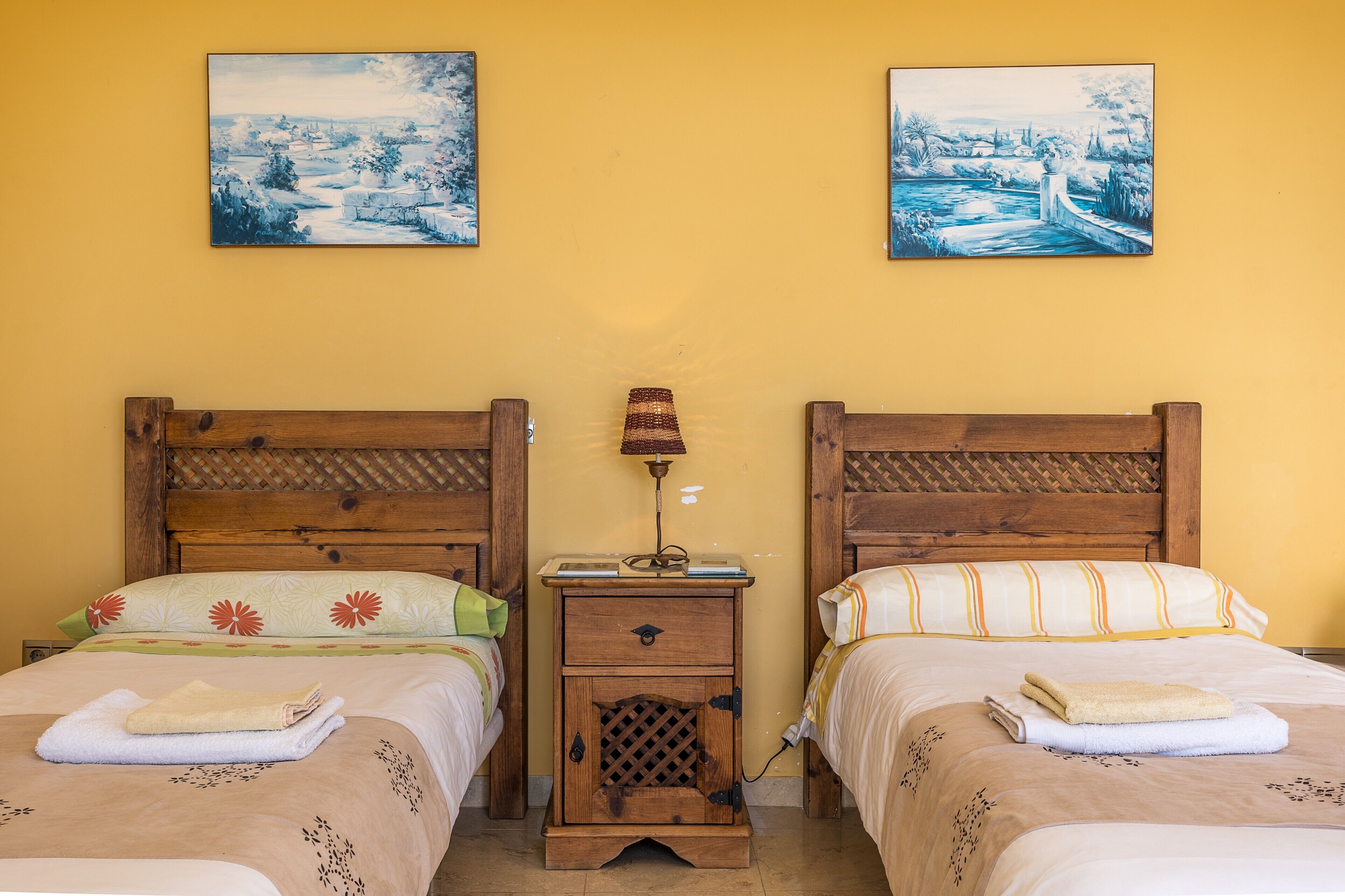 Una habitación doble con dos camas de madera, una mesita de noche con lámpara y dos cuadros en la pared.
