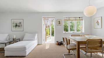 Una habitación moderna y luminosa con un sofá blanco, una mesa de comedor con sillas y grandes ventanales con vistas a zonas verdes.