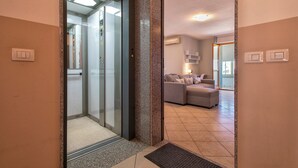 Un ascensor con puerta de cristal, una sala de estar con sofá y una mesa de centro, y una unidad de aire acondicionado instalada en la pared.
