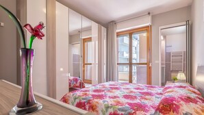 Una habitación de hotel con cama, espejo, un jarrón con flores y ventana con cortinas.