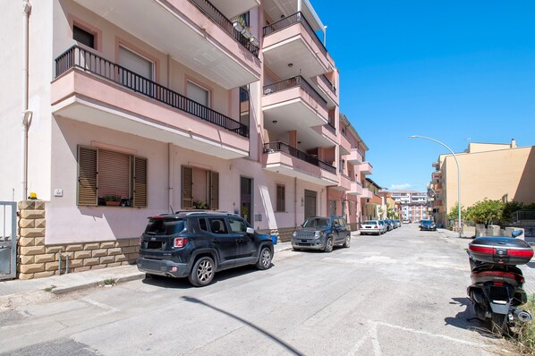 Un immeuble résidentiel à plusieurs étages, doté de balcons, avec des voitures garées devant et un ciel bleu dégagé.