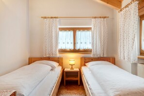 A room with two single beds, a nightstand with a lamp, a window with curtains, and wooden walls.
