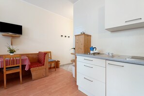 Een kleine keuken met witte kastjes, een houten eettafel met een rood-wit geruit tafelkleed, en een ingebouwde televisie.
