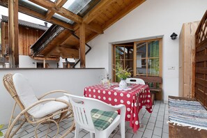 En overdækket terrasse med fletstole, et bord med rødt prikket dug og en træbænk.