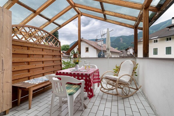 A covered patio with a wooden bench, a table with a red tablecloth, and a wicker chair.
