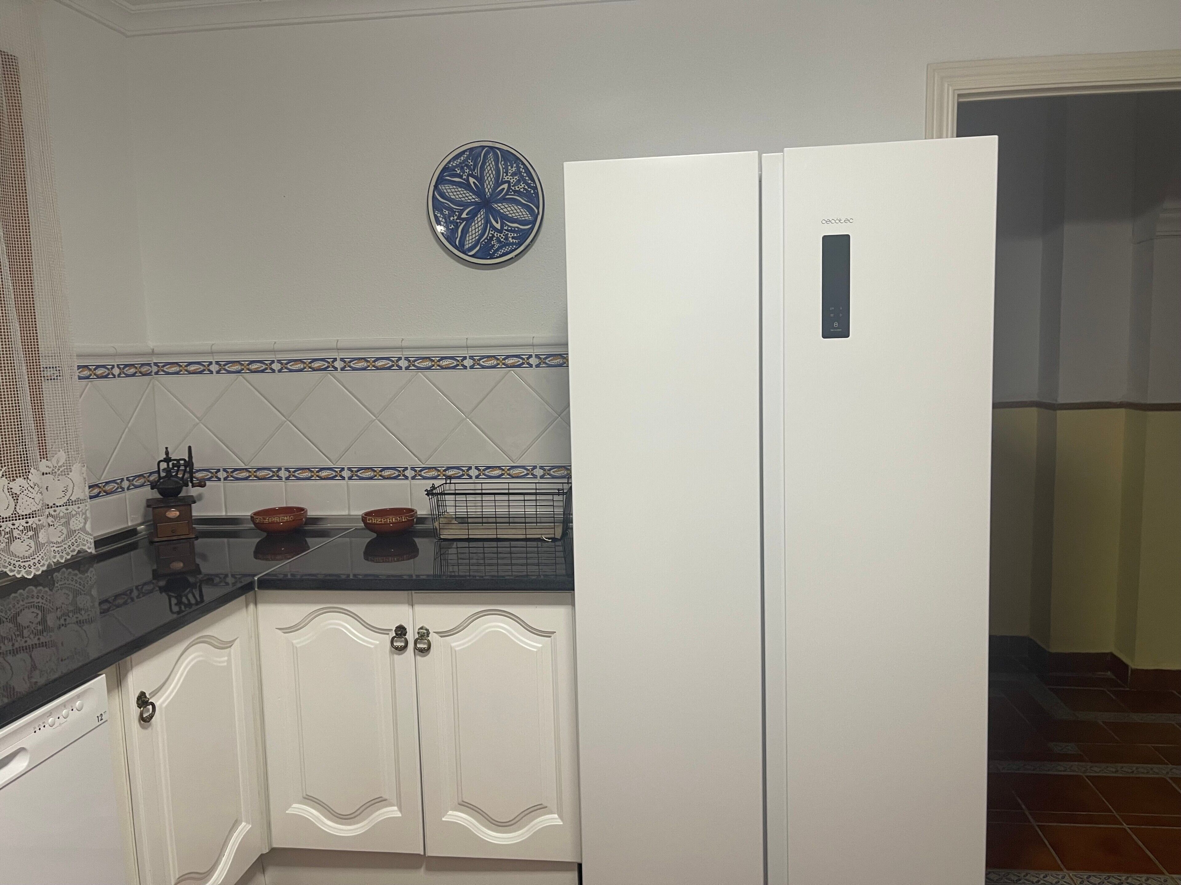 Una cuina amb armaris blancs, tauleig negre, plat decoratiu blau i frigorífic blanc.