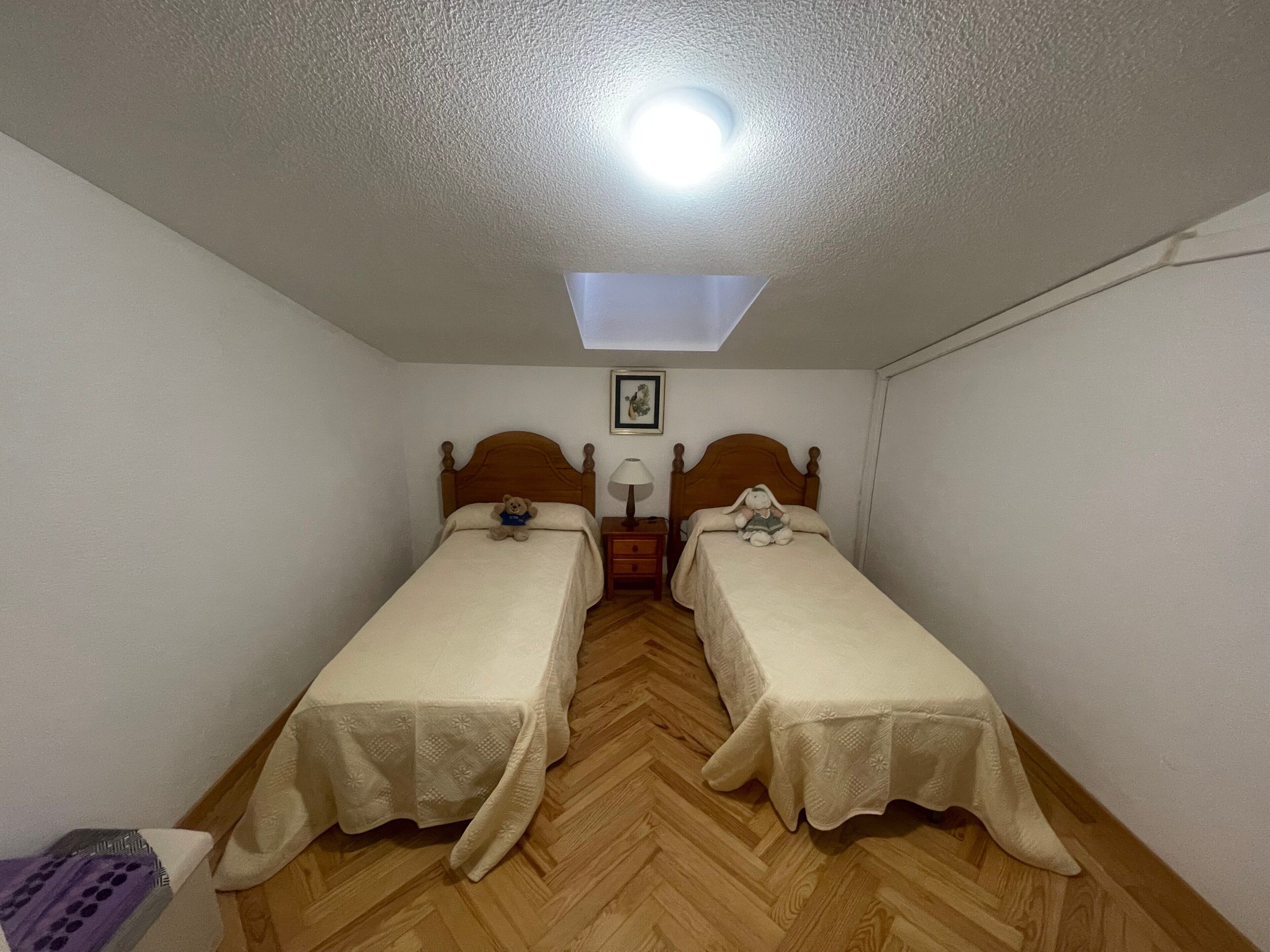 Ein kleines Zimmer mit zwei Einzelbetten, einem Nachttisch mit Lampe, einer Deckenleuchte und einem gerahmten Bild an der Wand.