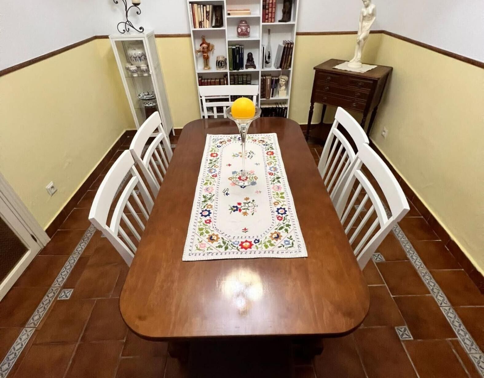 Una mesa de comedor con camino de mesa floral, sillas blancas, un florero con una flor amarilla y una estantería al fondo.