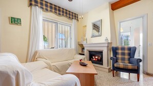Ein Wohnzimmer mit einer Couch, einem Holztisch, einem Karosessel, einem Kamin und Blick durch ein Fenster.