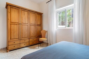 Quarto com guarda-roupa de madeira, cadeira de vime, cama com colcha azul e janela com cortinas.