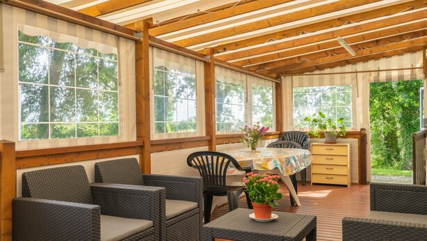 Eine überdachte Terrasse mit Korbmöbeln, einem Tisch mit Blumenmuster und Kübelpflanzen.