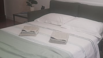 Una cama bien hecha con sábanas blancas y una manta verde, una mesita de noche con una lámpara y una planta en maceta en la pared.