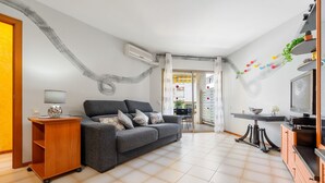 A living room with a sofa, a wooden side table, a TV, and a kitchen area with cabinets. - Apartment 'Bumbai' with Shared Pool, Private Garden and Air Conditioning (Creixell)