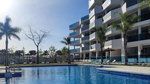 A swimming pool with a clear blue water, surrounded by a modern building with multiple balconies and palm trees. - Apartment 'Jardines De Las Lagunas' with Mountain View, Wi-Fi and Air Conditioning (Las Lagunas de Mijas)