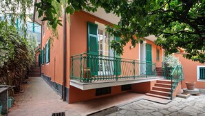 A building with a stone-paved courtyard, a balcony with green railings, and a staircase leading to the entrance.