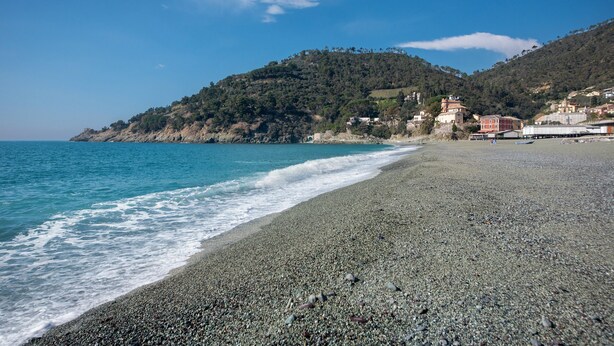 A pebble beach with a view of the sea and a hillside with buildings.
