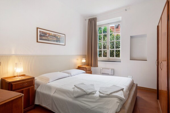 A hotel room with a bed, bedside tables, a window with curtains, a radiator, and a framed picture on the wall.