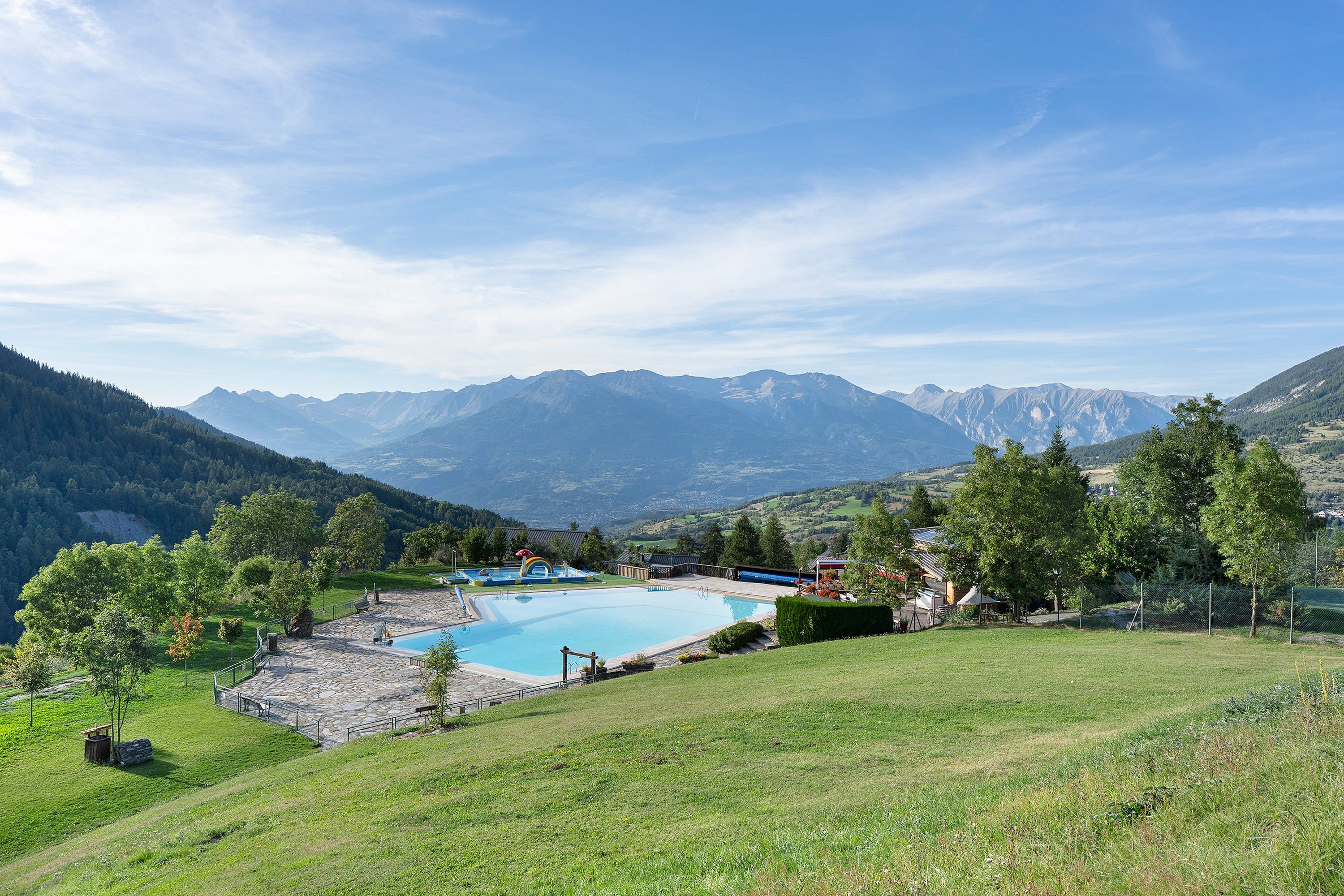 A swimming pool with a stone deck, surrounded by a grassy area and trees, with mountains in the background.