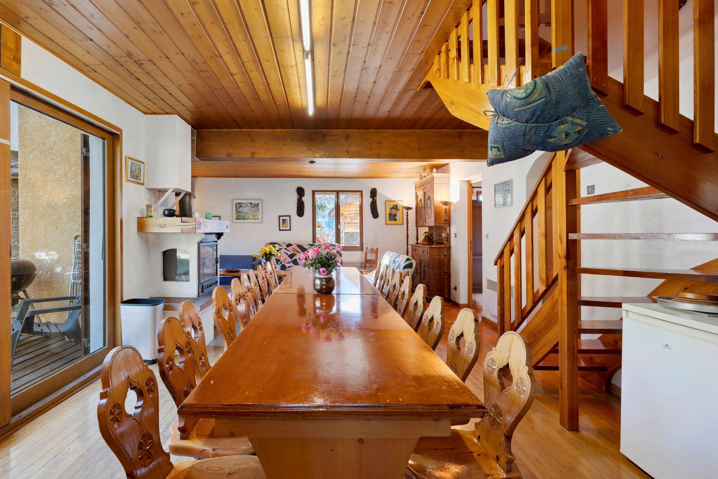 Uma sala de jantar ampla, com uma longa mesa de madeira rodeada por cadeiras de madeira. Uma escadaria com corrimão de madeira leva a um andar superior.
