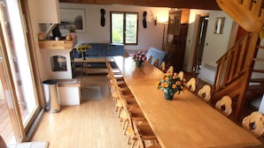 Una larga mesa de comedor de madera con sillas, un sofá azul y un florero en una cocina con suelo de madera y vistas al exterior.