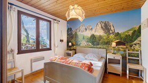 Un dormitorio con cama, ventana, lámpara de pared y un mural con paisaje montañoso.