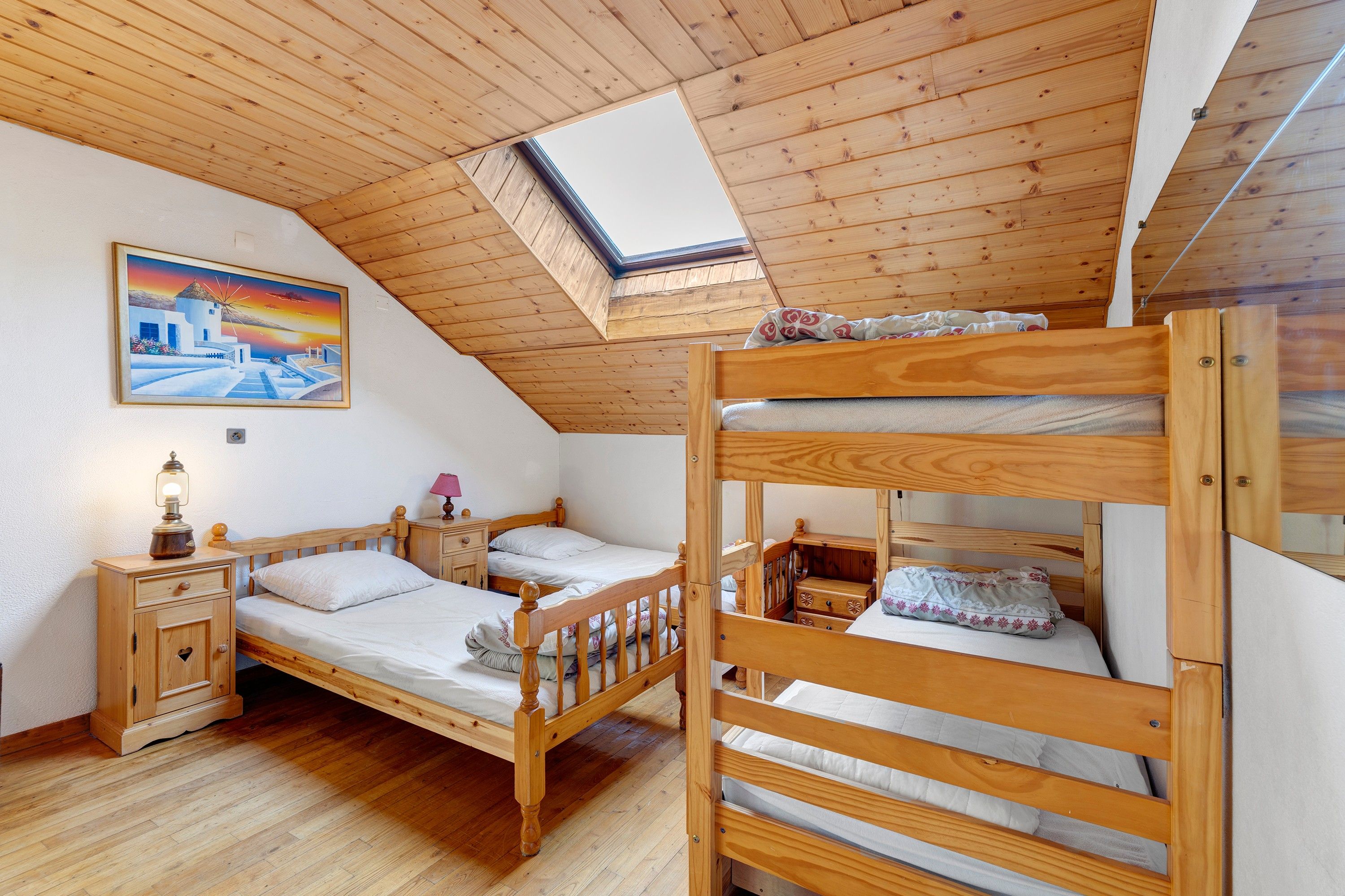 Une pièce avec un plafond en pente, des lits superposés en bois, une table de chevet avec une lampe, une commode et un tableau représentant un paysage côtier.