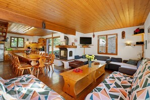 Amplia sala de estar con techo de madera, una mesa de comedor con sillas, un conjunto de sofás, un televisor y una chimenea.