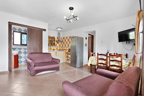 A living room with a sofa, a dining area with a table and chairs, a kitchen with a refrigerator, and a TV mounted on the wall.