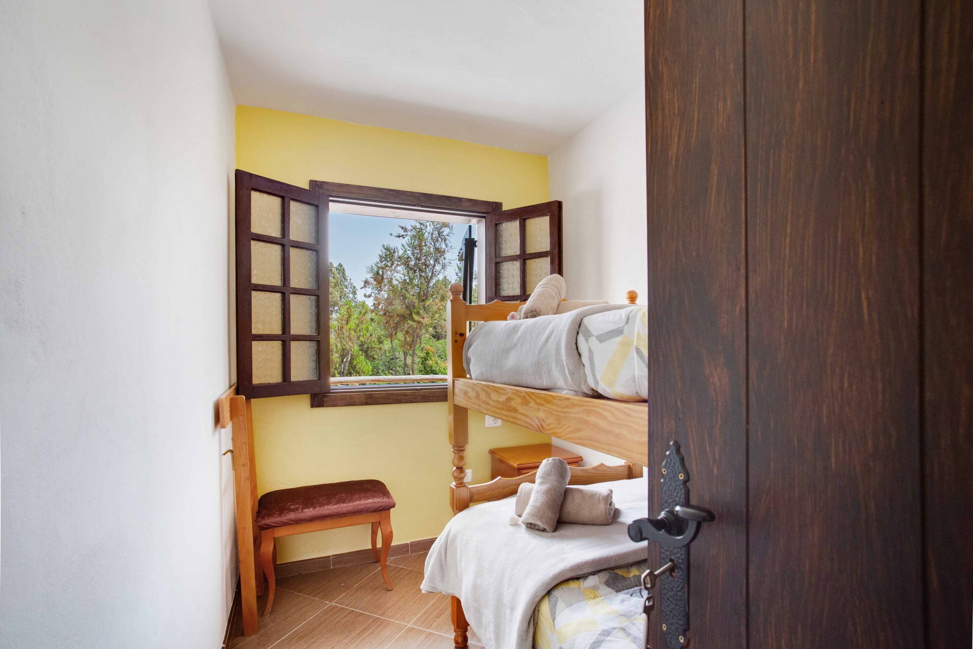A room with a bunk bed, a bench, a window with open shutters, and a door.