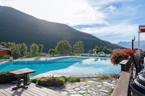 Une piscine aux eaux cristallines, bordée d’une terrasse en bois, entourée de verdure et avec des montagnes en toile de fond.