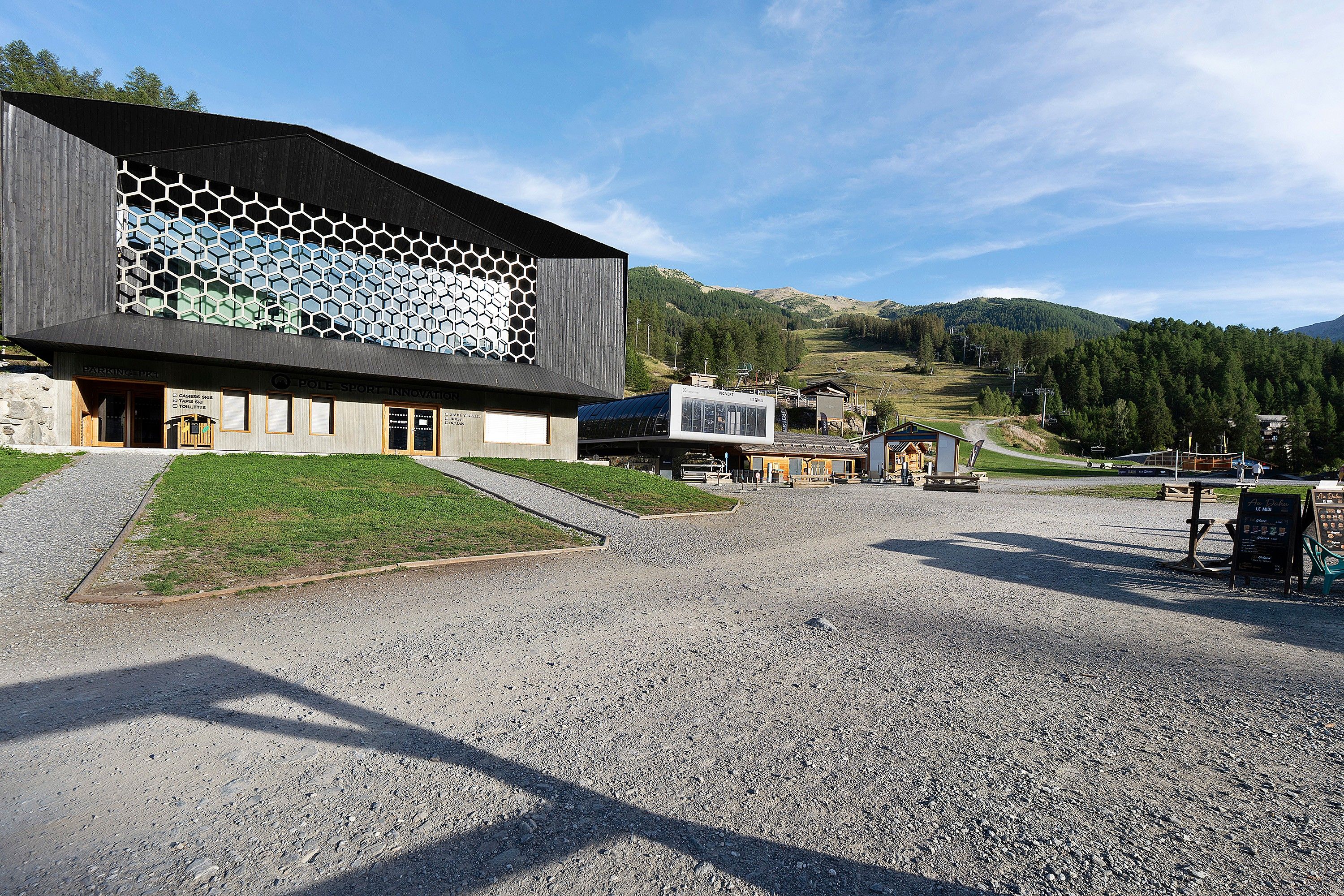 Un bâtiment moderne à la façade hexagonale, doté d’un parking en gravier et s’inscrivant dans un paysage montagneux.
