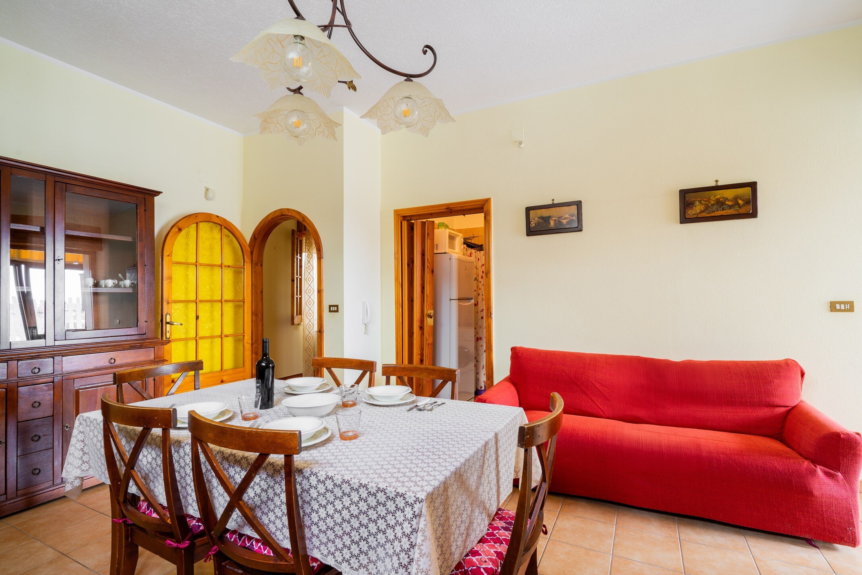 Un comedor con una mesa preparada para cuatro, sillas de madera, un sofá rojo y un mueble con puertas de vidrio.
