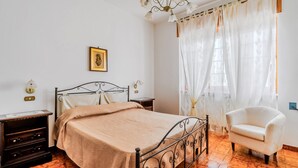Une chambre à coucher avec un lit, une chaise, une table de chevet, une lampe et une fenêtre avec des rideaux.