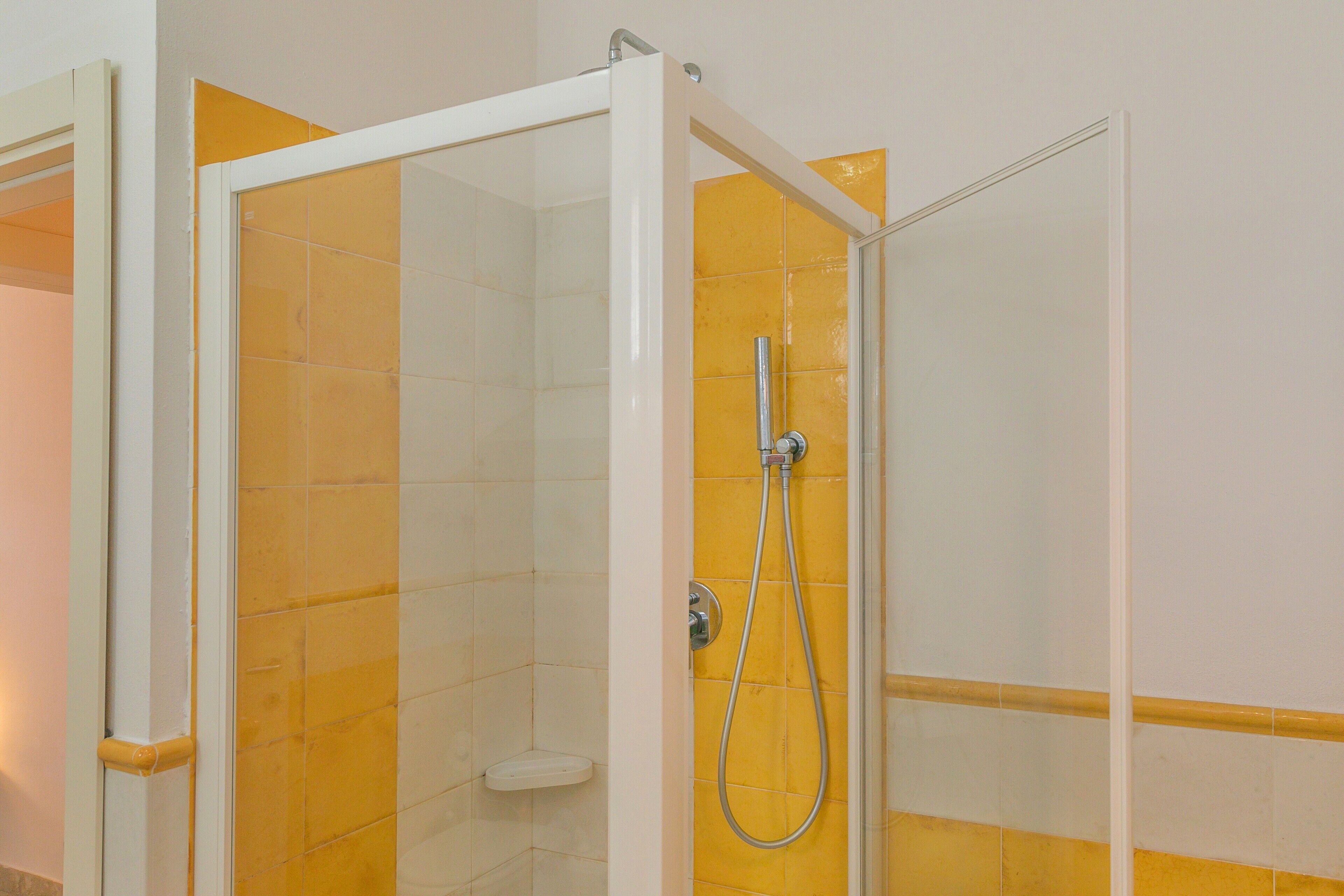 A shower with a glass door, yellow and white tiles, and a handheld showerhead.