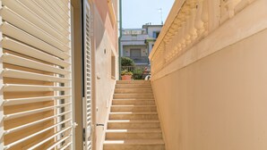 Un escalier étroit menant à un balcon doté d’une balustrade, une porte avec des stores et une vue sur les bâtiments adjacents.