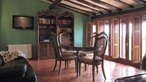 A room with a wooden ceiling, a television mounted on the wall, a bookshelf, a dining table with chairs, and a radiator. - La Tahona: Sustainable Country House for Cyclists, Starlight-Certified in Lerín (Lerín)