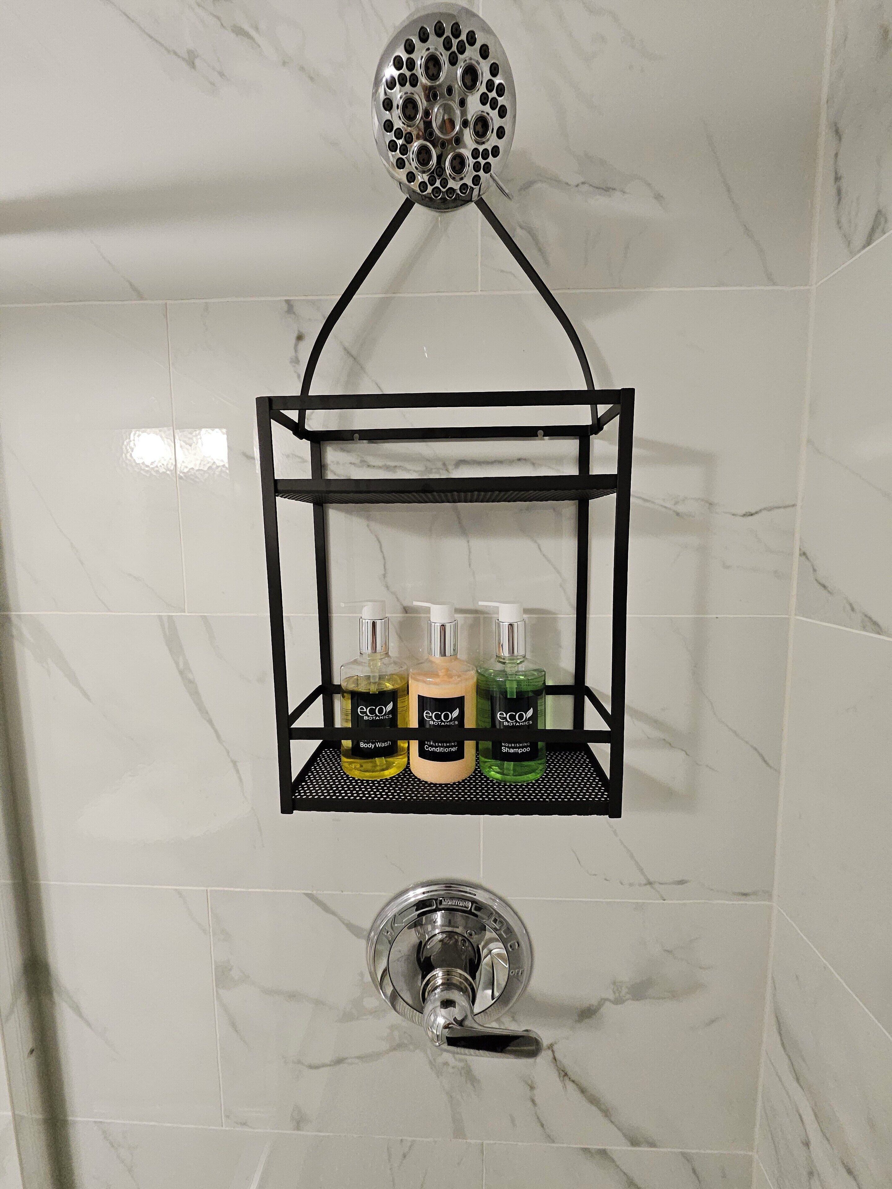 Un baño con un estante de metal negro que contiene tres botellas de artículos de aseo ecológicos, un cabezal de ducha y un grifo cromado.