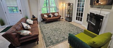 Una sala de estar con un sofá marrón, un sillón verde, un tapete estampado, una chimenea y un televisor montado en la pared.