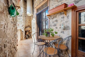 En terrasse med stenvægge, et rundt træbord og metalstole, en grøn vandkande og et vindue med en træhylde over.