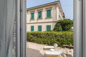 Un bâtiment à la façade symétrique, doté de volets verts et d’une terrasse en gravier agrémentée de chaises longues blanches.