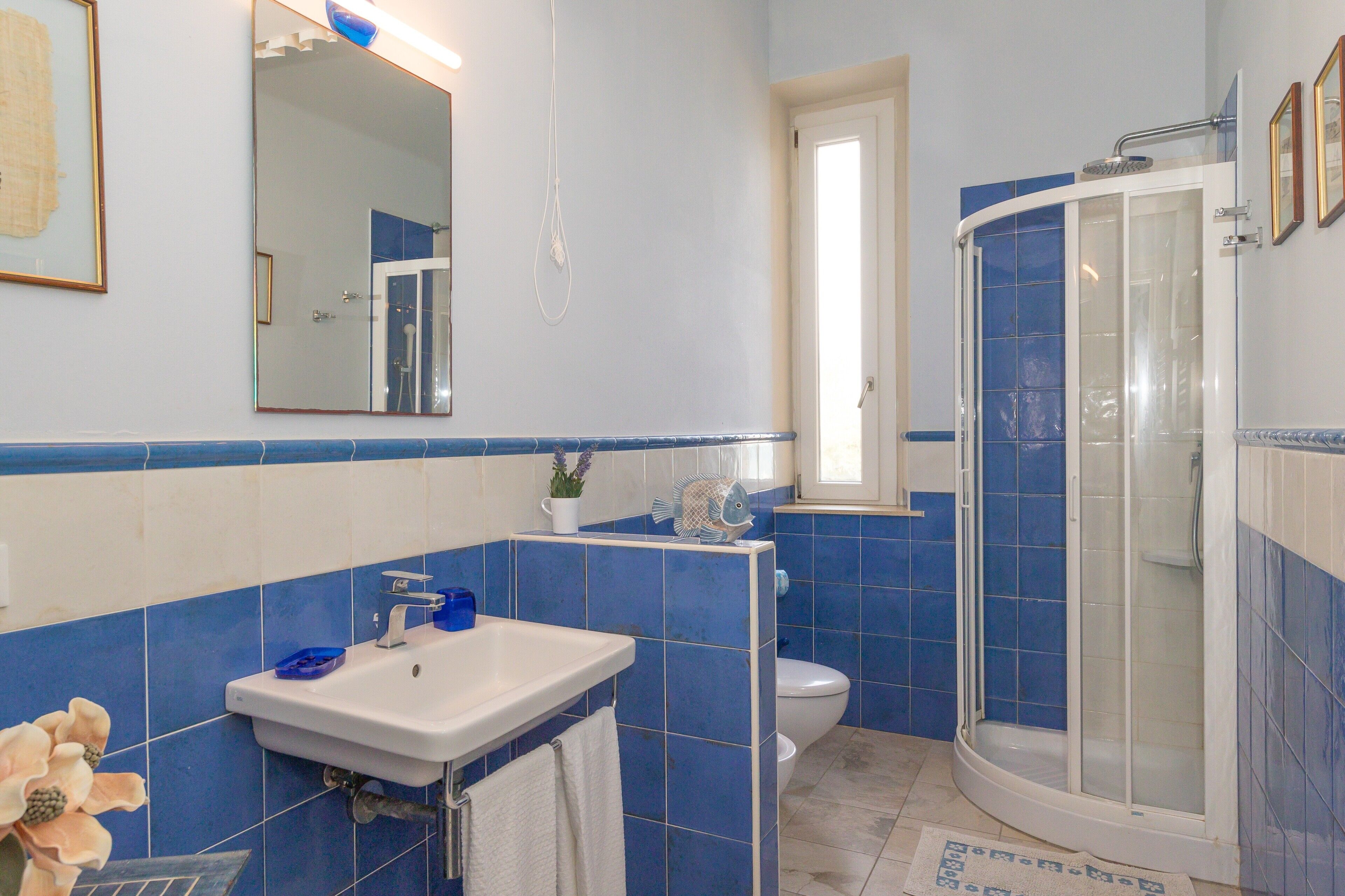 Un baño con lavamanos blanco, azulejos azules, cabina de ducha de vidrio y una ventana pequeña.