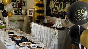 Una larga mesa preparada para una celebración con temática del 50 aniversario, incluyendo globos, un pastel y diferentes platillos.