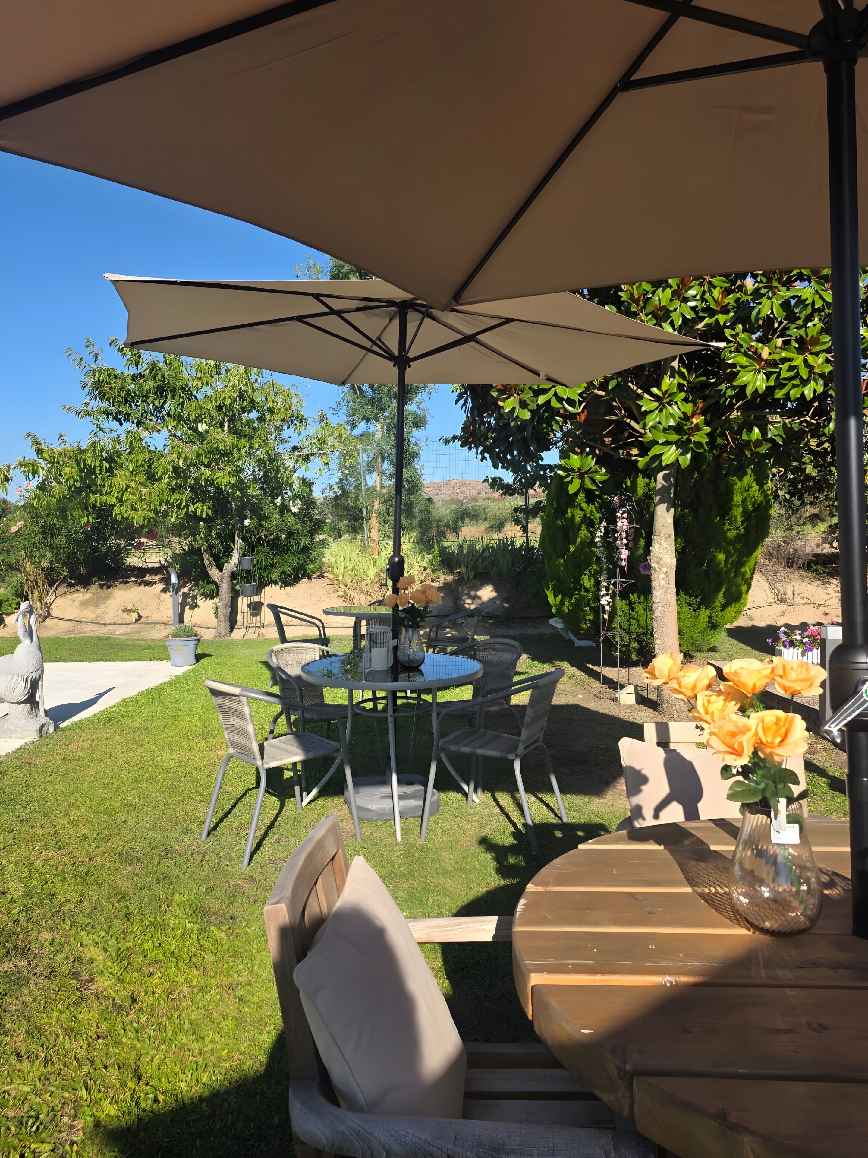 An outdoor patio with a table and chairs under a large umbrella, surrounded by greenery and a statue.