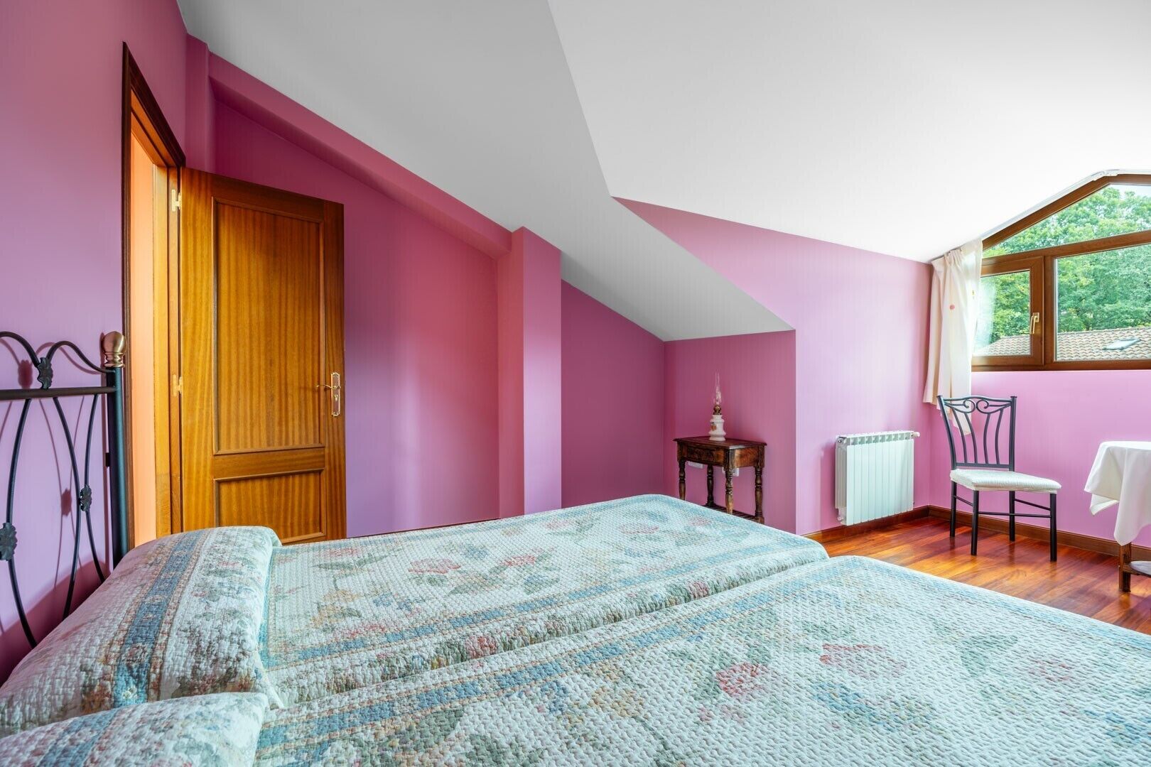 A bedroom with pink walls, a wooden door, a bed with a patterned bedspread, a small table with a lamp, a chair, and a window with a view of greenery.