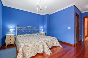 Ein Schlafzimmer mit blauer Wand, Holzboden, einem Bett mit floralem Überwurf, zwei Nachttischen mit Lampen und einer Deckenleuchte.