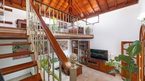 Un salón amplio con una escalera de madera, un balcón, un televisor y varias plantas.