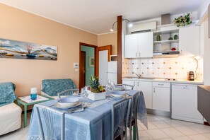 A dining area with a table set for four, a sofa, a small side table with a candle, and a kitchen area with cabinets and a dishwasher. - Apartment 'Casa Vacanza Marina Di Pietrasanta' with Mountain View, Wi-Fi and Air Conditioning (Pietrasanta)